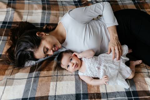 Mãe deitada de bruços na colcha xadrez observando carinhosamente a filha.'