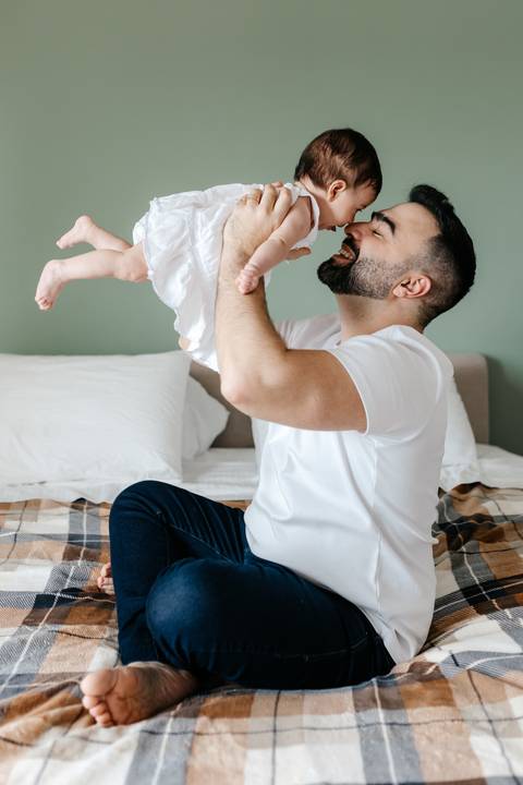 Pai erguendo a bebê no ar com os braços esticados enquanto a mãe observa sorrindo.'
