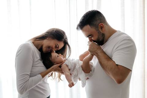 Mãe segurando a bebê no colo enquanto o pai beija o pezinho dela em frente à janela.'