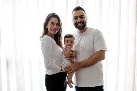Retrato oficial dos pais em pé sorrindo com a bebê em frente à cortina iluminada.'