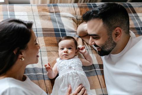 Pais deitados de bruços na colcha xadrez sorrindo e conversando com a filha.'