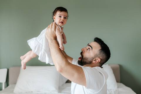 Pai em pé no quarto erguendo a bebê no ar e olhando para ela com admiração.'