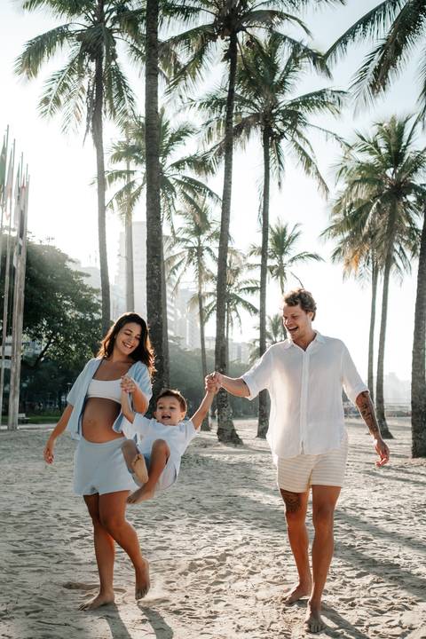 Família sorridente caminhando de mãos dadas na areia da praia entre coqueiros.'