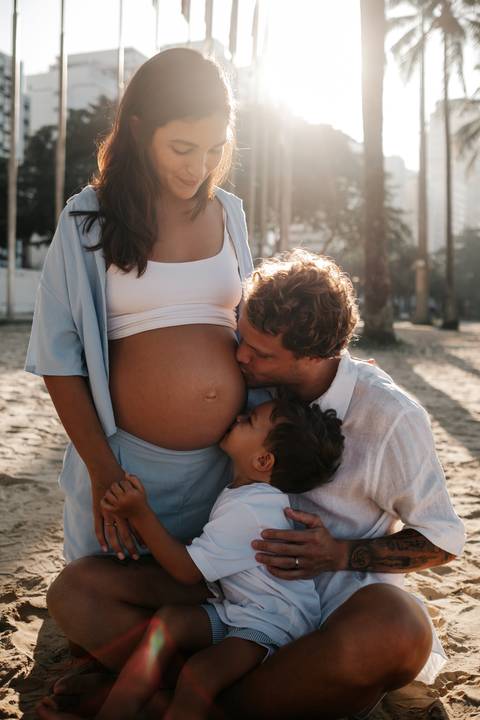 Close do pai e do filho pequeno beijando juntos a barriga exposta da gestante.'