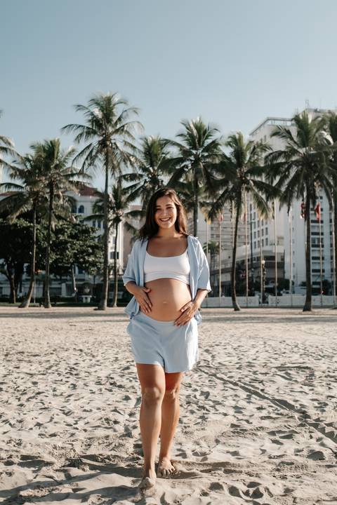 Retrato frontal da gestante com a barriga à mostra sorrindo em frente aos coqueiros.'
