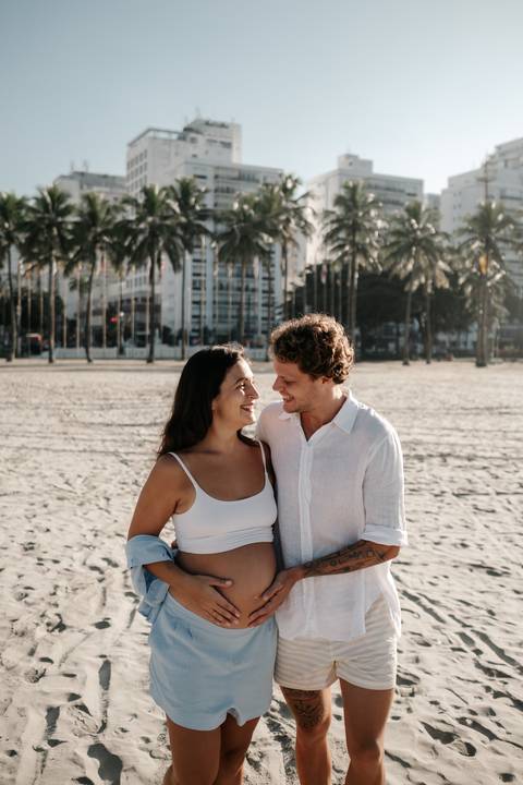 Casal abraçado na areia trocando olhares apaixonados com prédios e coqueiros ao fundo.'