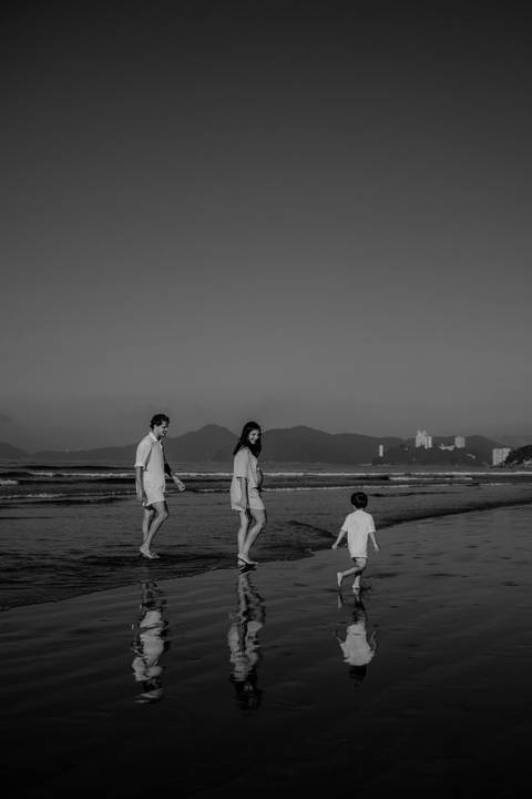 Versão em preto e branco da família caminhando de costas à beira-mar.'