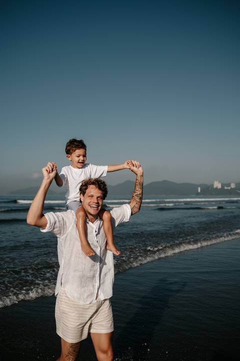 Pai sorrindo carregando o filho pequeno nos ombros na beira da praia.'