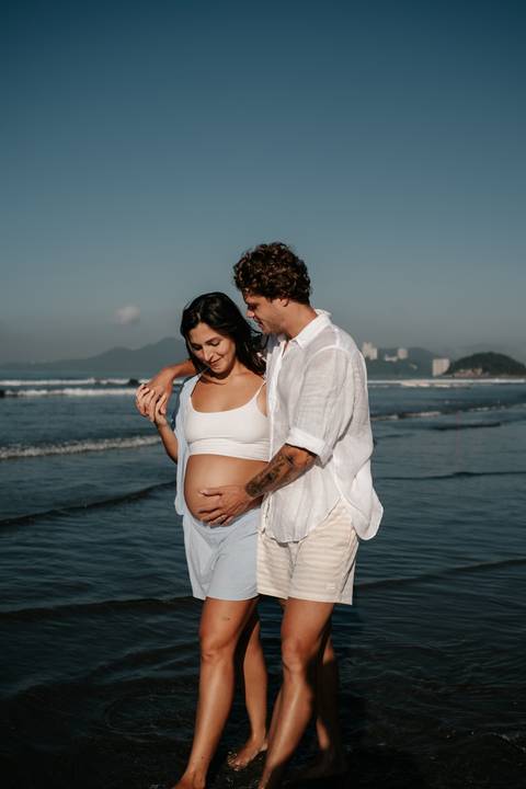 Casal abraçado olhando para a barriga exposta da gestante na praia.'