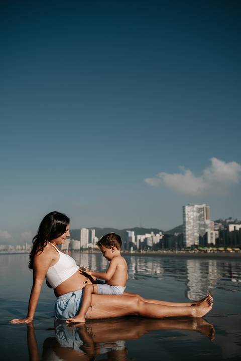 Perfil da mãe e do filho com os prédios da cidade ao fundo na praia.'