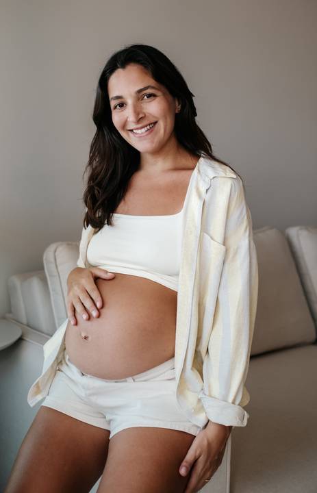 Close da gestante sentada no sofá em ambiente interno, com camisa aberta mostrando a barriga.'