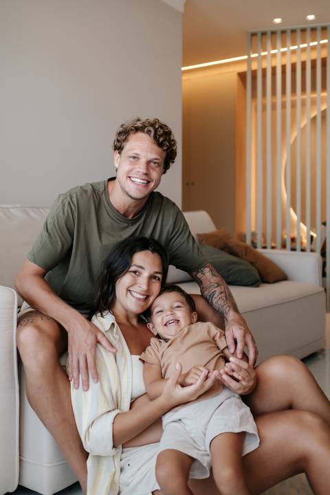 Retrato frontal da família sorrindo e reunida no chão ao lado do sofá.'