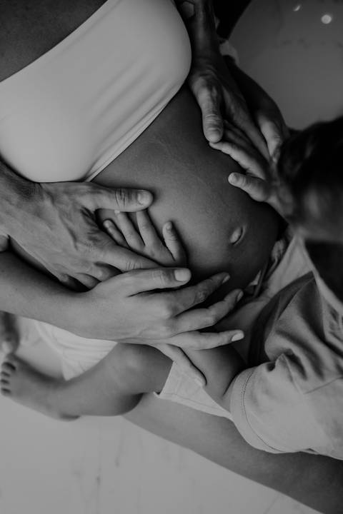 Versão em preto e branco do close das mãos do pai e do filho na barriga da gestante.'