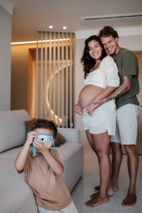 Filho pequeno em primeiro plano brincando com uma câmera fotográfica de brinquedo azul enquanto os pais posam ao fundo.'