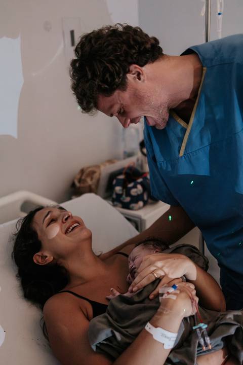 Companheiro sorrindo e comemorando ao olhar para a mãe e o bebê recém-nascido na cama.'