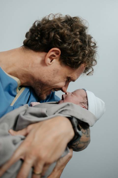 Pai sorrindo e olhando bem de perto para o rostinho do bebê recém-nascido.'