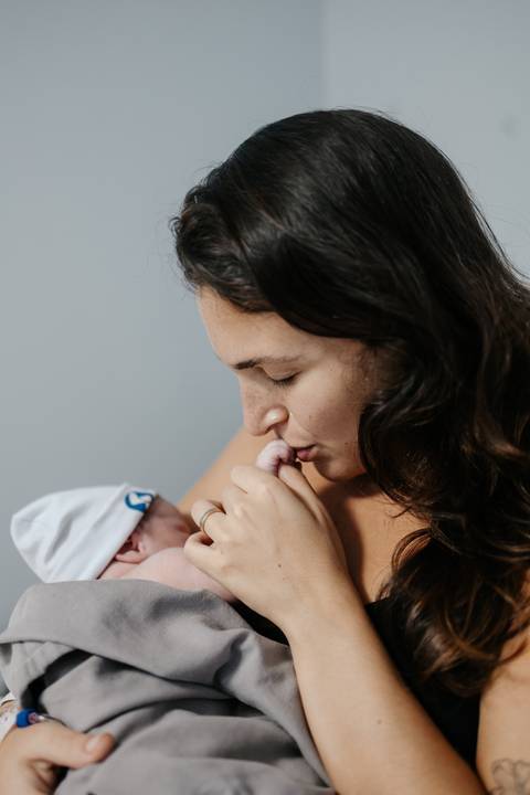 Mãe inclinada aproximando o rosto e beijando a mãozinha de seu bebê.'