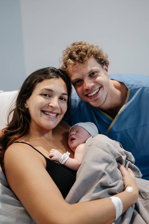 Retrato do casal sorridente posando com o bebê recém-nascido na cama.'
