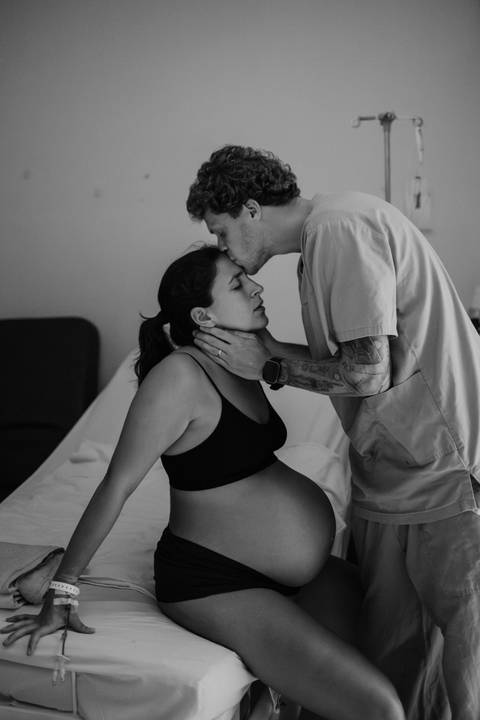 Versão em preto e branco do companheiro beijando a testa da gestante na cama.'