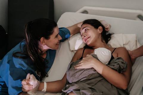 Mãe deitada com o bebê no colo recebendo o carinho e o olhar sorridente da profissional de saúde.'