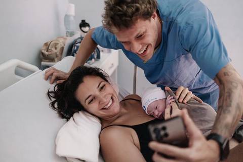 Casal sorridente tirando uma foto de celular (selfie) com o bebê deitado no peito da mãe.'