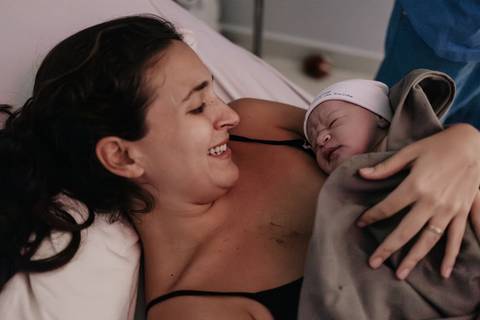 Mãe sorrindo e olhando com ternura para o bebê recém-nascido deitado em seu colo.'