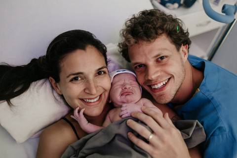 Retrato frontal do casal sorridente posando juntos com o bebê na cama hospitalar.'