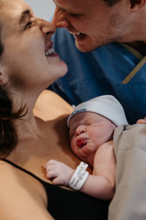 Registro aproximado do bebê dormindo serenamente no peito da mãe enquanto os pais se aninham.'
