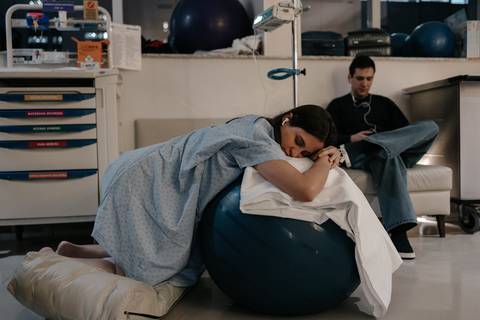 Gestante debruçada sobre a bola de pilates no quarto de hospital, com o companheiro ao fundo.'