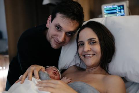 Casal sorridente na cama de hospital segurando o recém-nascido no colo.'