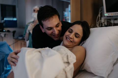 Casal sorridente e aliviado na cama hospitalar com o bebê recém-nascido nos braços.'