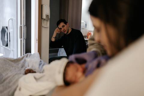 Mãe deitada com o bebê no peito enquanto o pai observa ao fundo no quarto do hospital.'