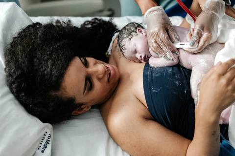Vista superior do recém-nascido no peito da mãe enquanto a equipe médica realiza os primeiros cuidados.'