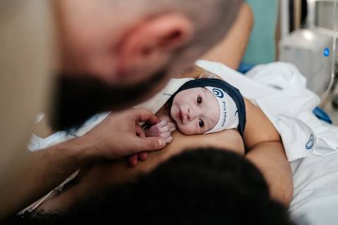 Mãos do pai e da mãe acolhendo o recém-nascido com touquinha durante o contato pele a pele.'