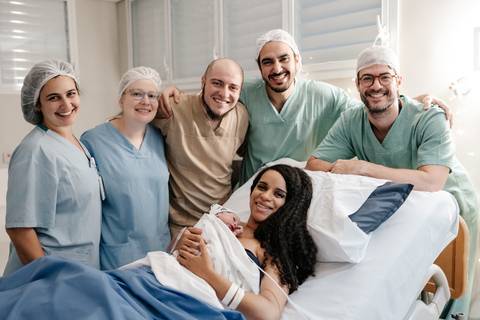 Foto em grupo da família sorridente na cama hospitalar acompanhada por toda a equipe médica.'