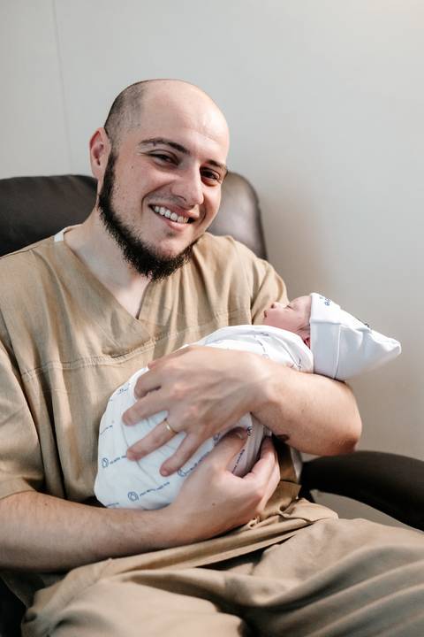 Pai sorridente segurando com cuidado o bebê recém-nascido envolto em uma manta nos braços.'
