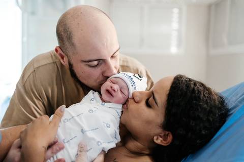 Pai e mãe beijando simultaneamente a cabeça do recém-nascido no colo.'