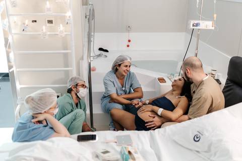 Visão ampla da gestante sentada na borda da cama cercada pelo parceiro e equipe de assistência.'