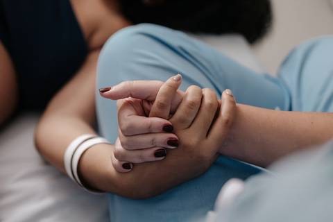 Mãos da enfermeira e do acompanhante segurando firmemente as mãos da gestante na cama.'