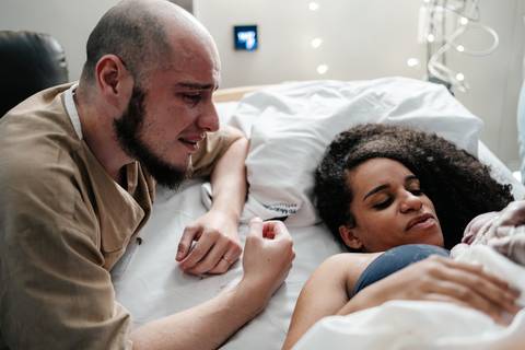 Pai chorando emocionado ao lado da mãe deitada na cama hospitalar com o recém-nascido.'