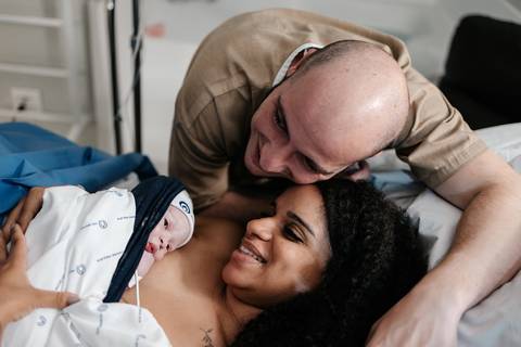 Casal sorridente na cama do hospital abraçando e olhando para o recém-nascido no peito da mãe.'