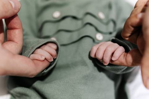Detalhe das mãos dos pais segurando delicadamente as mãozinhas do bebê.'