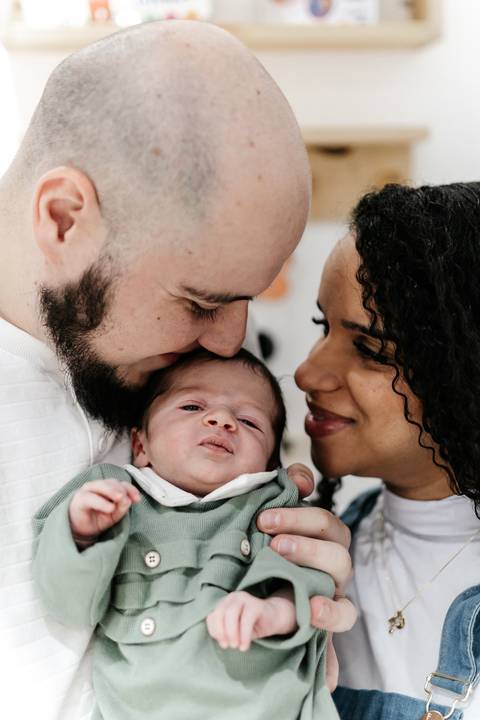 Pai e mãe de rostos colados beijando o topo da cabeça do bebê no colo.'
