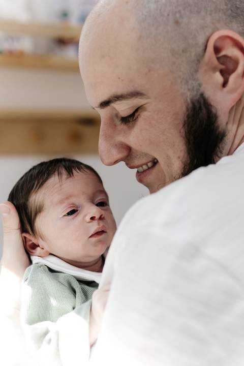 Pai sorrindo de perto para o bebê recém-nascido que segura em seus braços.'