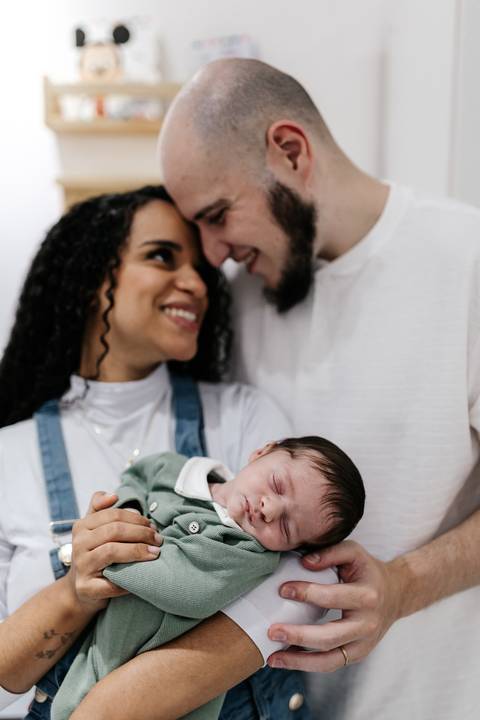 Casal sorrindo e trocando olhares amorosos enquanto seguram o bebê dormindo.'