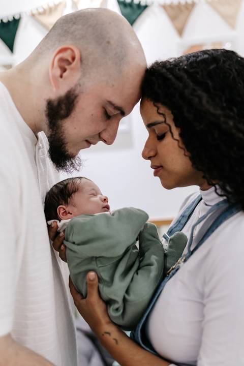 Casal de olhos fechados e testas encostadas segurando o bebê adormecido no colo.'