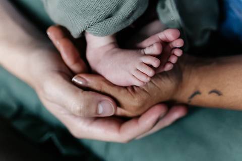 Mãos do pai e da mãe entrelaçadas envolvendo os pezinhos do bebê.'