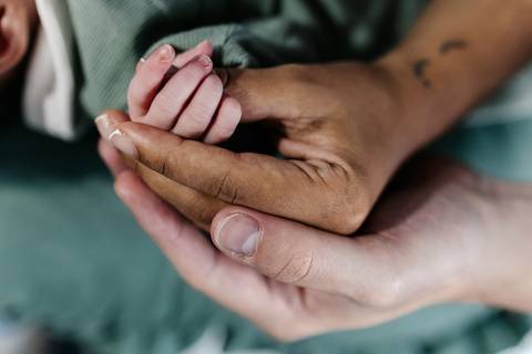 Mão do pai e da mãe juntas segurando com delicadeza a mãozinha do bebê.'