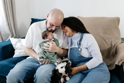Casal sorrindo no sofá com o bebê no colo e o pet da família ao lado.'