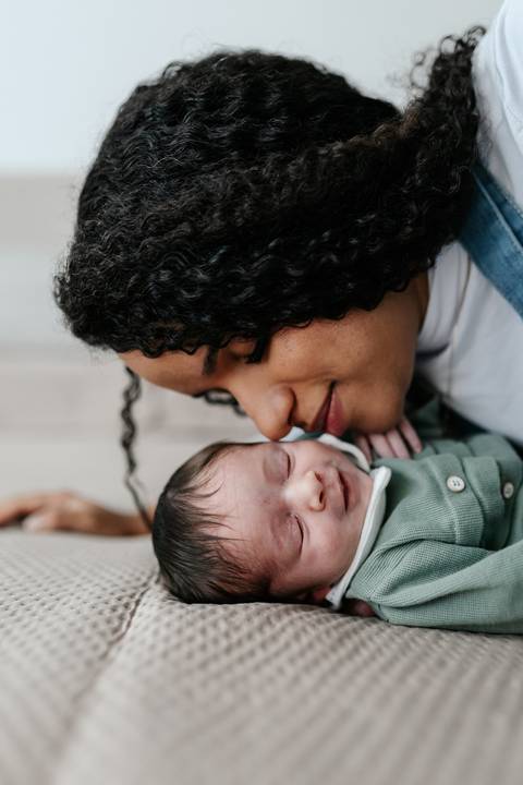 Mãe deitada ao lado do bebê adormecido observando-o com carinho.'